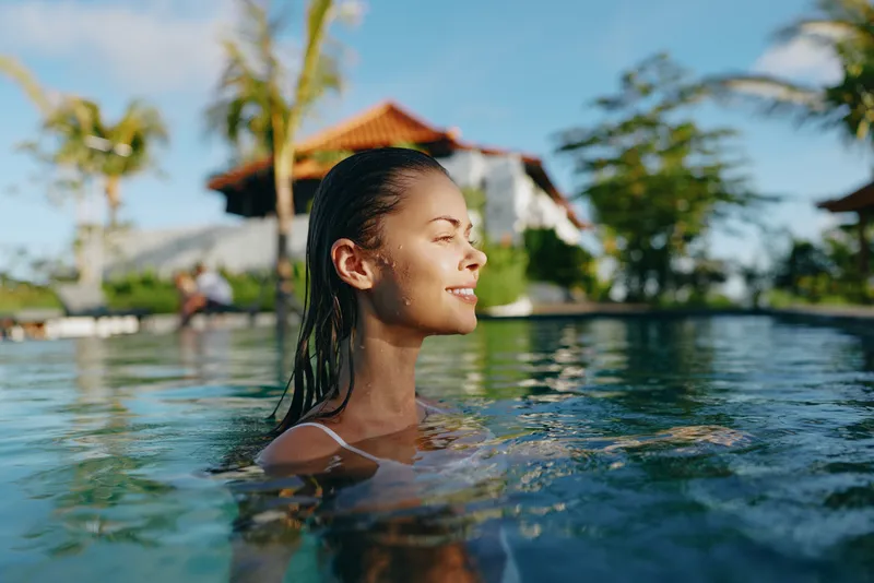 Bright woman enjoys a sunny pool moment, eyes closed, contented smile, warm breeze, clear water, tropical resort vibe, relaxed posture, summer leisure, vibrant colors, carefree lifestyle, outdoor