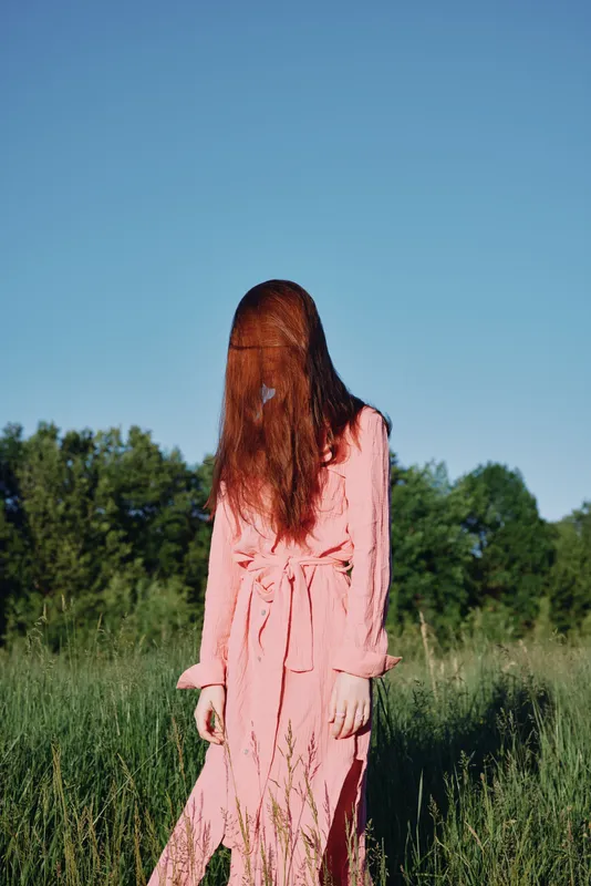 A young woman in a flowing pink dress with long red hair stands in a grassy field, exuding a sense of mystery and whimsy under a clear blue sky