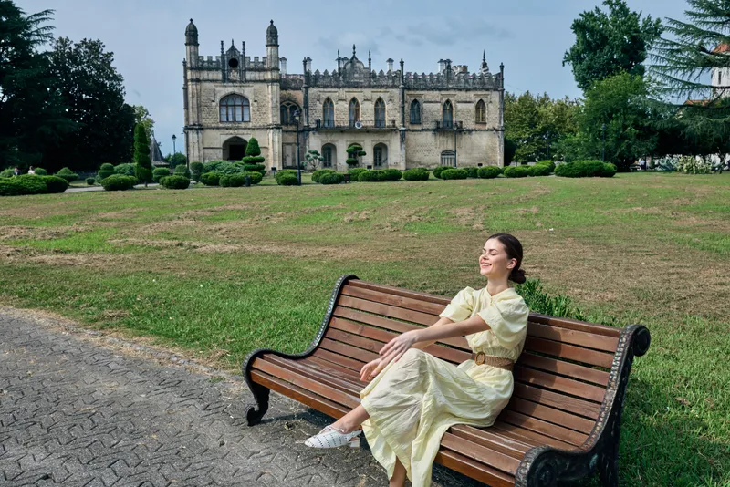 Woman in a light yellow dress sits on a wooden bench in a park, with a historic castle in the background, enjoying a peaceful moment outdoors today.