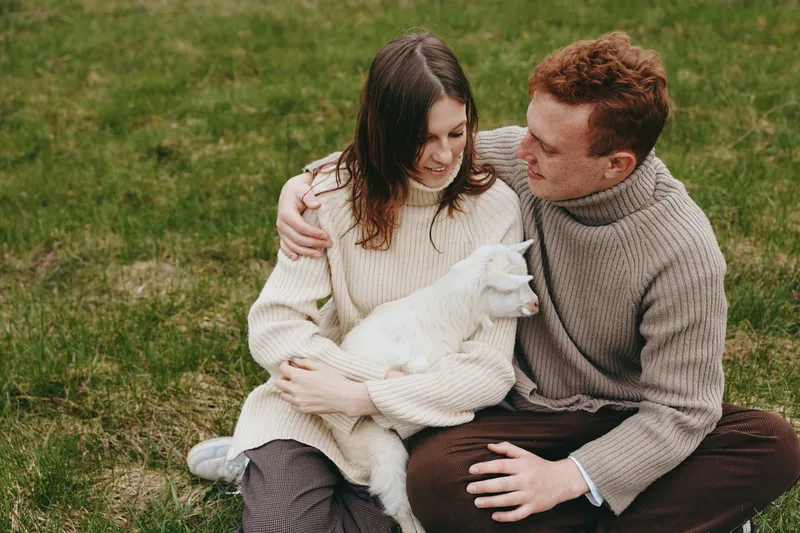 Couple sitting on grass in cozy sweaters, embracing a white goat kid, sharing affection outdoors in a peaceful rural setting, expressing warm companionship and gentle bonding