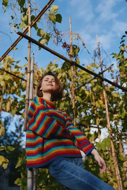 Woman in rainbow sweater, portrait in garden by wooden trellis, casual jeans and sunlight, autumn foliage and relaxed pose looking up for outdoor lifestyle and seasonal mood