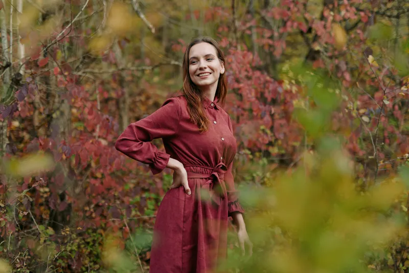 A candid portrait of a smiling woman in a burgundy dress standing in a vivid autumn scene, conveying authenticity and credibility through natural pose and light.
