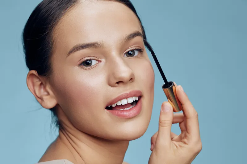 Young woman applying mascara with a joyful expression against a light blue background, representing beauty and cosmetics, perfect for makeup ads