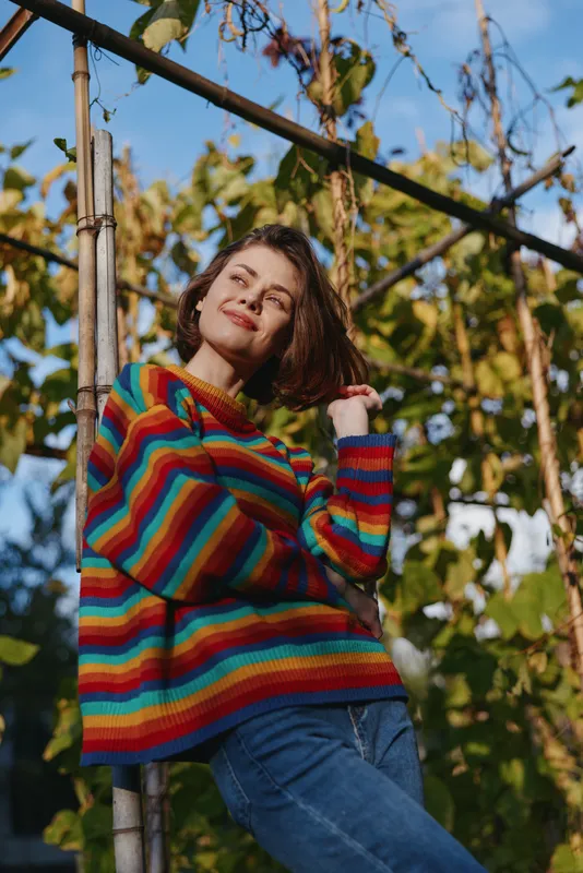 Woman in a rainbow sweater smiles in a garden portrait, casual pose among vine trellis and foliage with natural sunlight, colorful knitwear, cozy look and cheerful youthful mood.