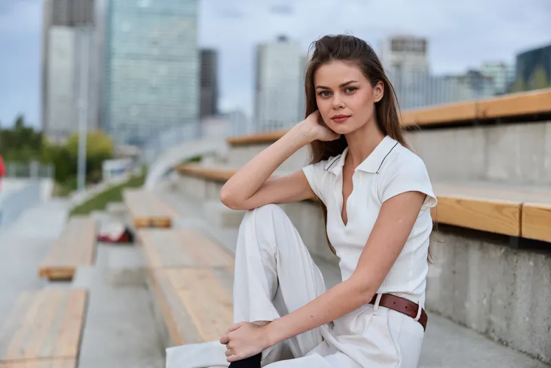 Young woman with confident expression sitting outdoors in casual white outfit in urban environment at daytime, city background, relaxed pose