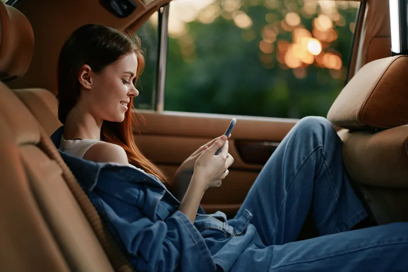 Relaxed young woman in casual denim outfit enjoying her smartphone while sitting in a luxury car during sunset.