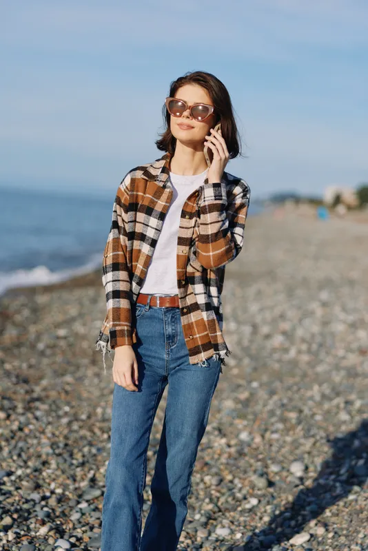 Woman on a sunny beach talks on her phone, wearing a plaid shirt, white tee and jeans. Casual coastal scene with clear sky, calm sea, and relaxed style for lifestyle and communication.