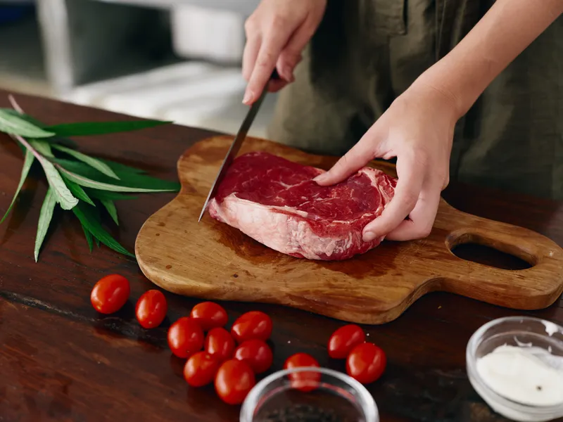 Женщина с ножом в руке нарезает свежее стейковое мясо для жарки на кухне; на столе соль, перец и другие специи, красные помидоры черри и зелень; она готовит ужин