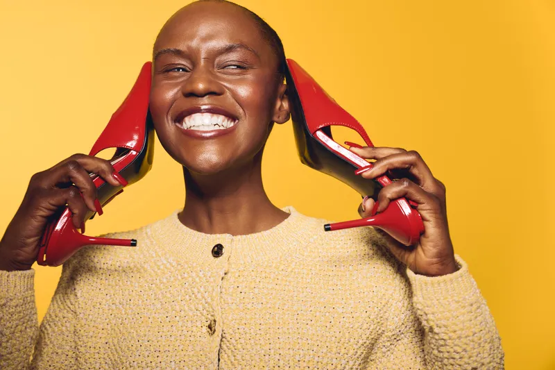 Happy smiling black woman with dark skin holds red high heels near face on yellow background. Concept of fashion, joy, beauty, lifestyle, and confident African ethnicity woman.