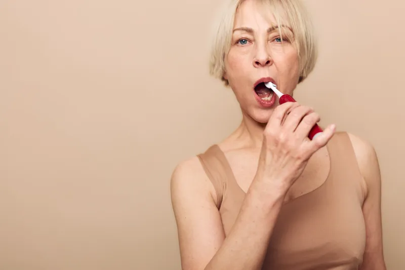 Blonde middle aged woman wearing beige tank top brushes teeth with electric toothbrush. Close up on neutral background. Concept of dental hygiene, morning routine, and self care habits.