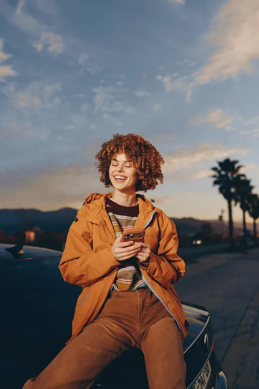 Cheerful woman lifestyle outdoors at sunset, curly hair smiling, casual jacket, holding a smartphone, relaxed posture on a quiet road, warm nature light in the evening breeze