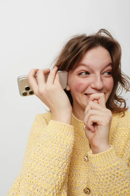 Young woman in yellow sweater holds smartphone near ear smiling shyly, looking aside. Concept of communication, happiness, casual lifestyle, and positive emotions in everyday life.
