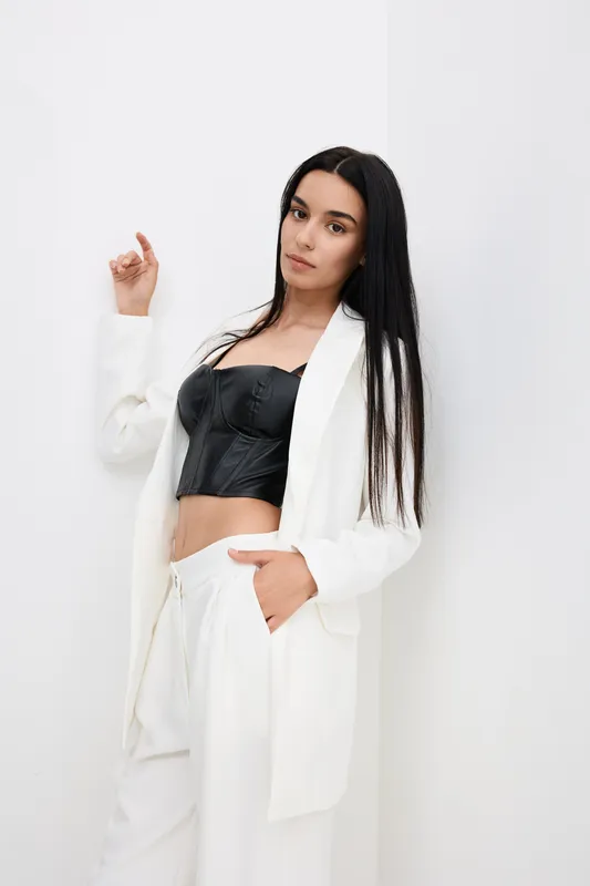 young woman in white suit with black corset top posing against white wall, long straight hair, stylish modern outfit, indoor minimalistic background