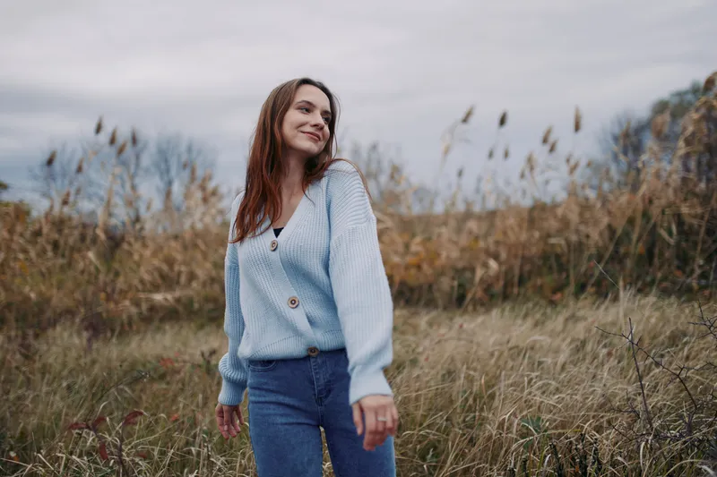 Young woman in a cozy blue cardigan walks through tall grass at dusk, photographed with natural light to convey authenticity and credibility in a candid, genuine lifestyle portrait.