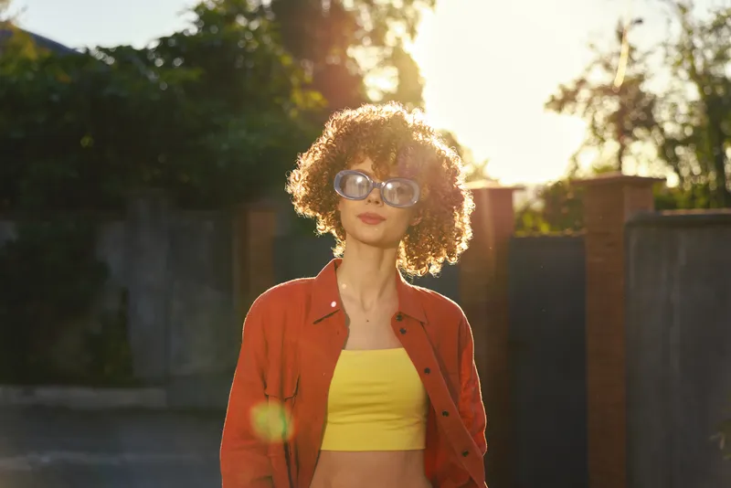 A stylish outdoor portrait of a woman in a yellow top and red jacket wearing sunglasses, captured in bright sunlight. A confident, casual lifestyle moment highlighting color and mood