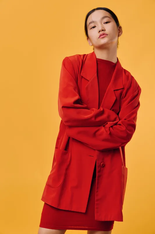 Young woman with serious expression wearing oversized red jacket and dress standing with crossed arms on yellow background. Fashion portrait with modern style and confident look.