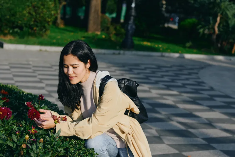 Woman kneels in a park, tending blossoms from a bush with careful touch, soft beige sweater, backpack, quiet outdoor moment shaped by sunlight, nature, and calm garden atmosphere