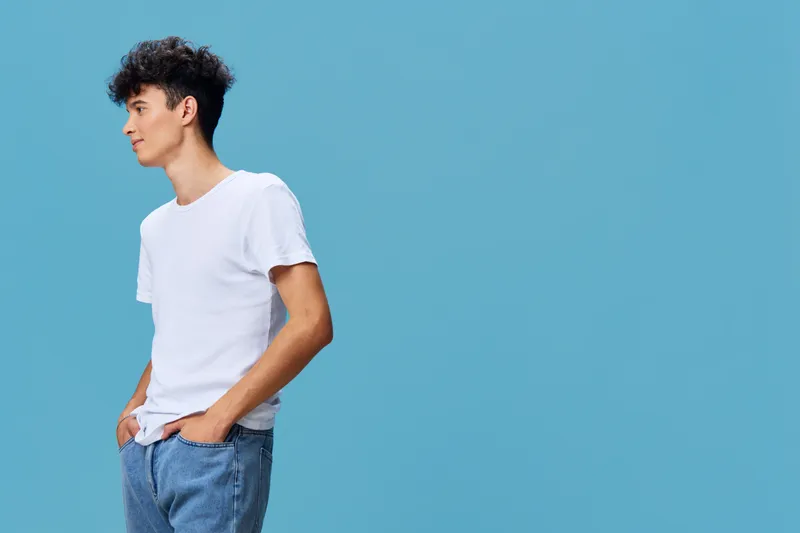 Young man wearing casual white t-shirt and blue jeans stands confidently with hands in pockets against solid light blue background, showing relaxed and thoughtful mood.