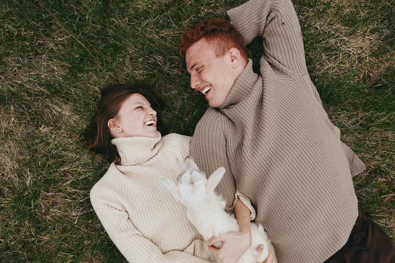A couple lying on grass in warm sweaters, smiling and embracing a fluffy rabbit, a cozy lifestyle scene outdoors with relaxed mood, playful affection, natural light, candid moment of love and comfort.