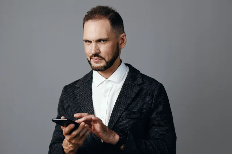 A poised business professional stands against a solid colored background, focused on his smartphone with a determined expression, projecting confidence, precision, and readiness for a corporate