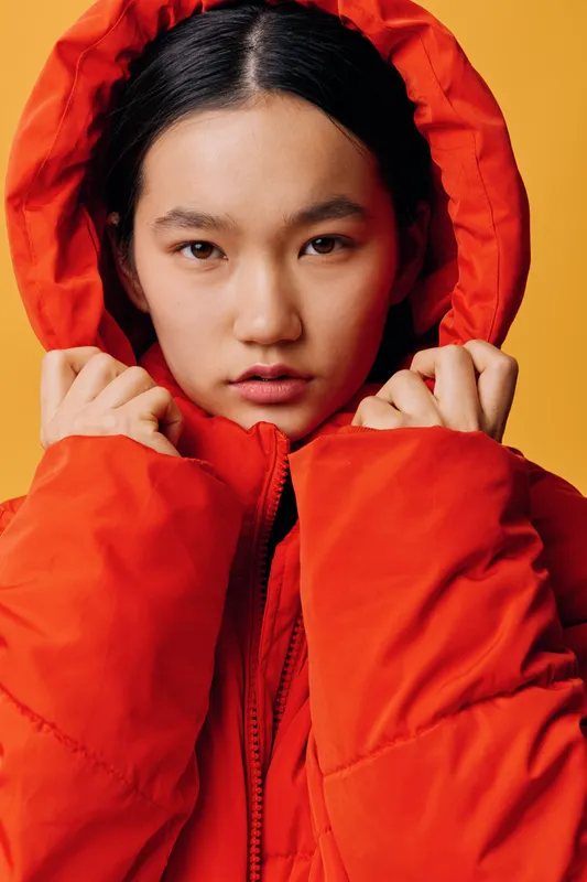 Young woman wearing bright orange puffer jacket with hood pulled over head, looking directly at camera with serious expression on solid warm background.