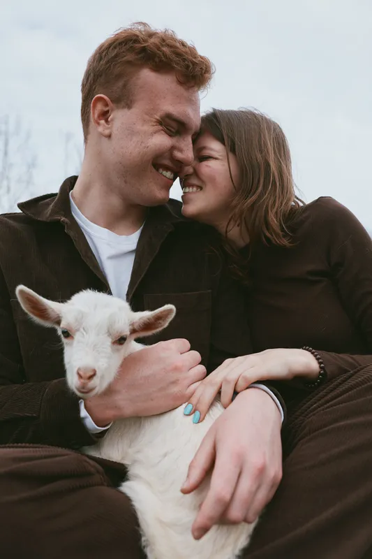 Joyful couple outdoors with a playful goat kid, embracing and smiling, sharing a warm candid moment in a sunlit rustic countryside setting their bond clear and sweet amid nature