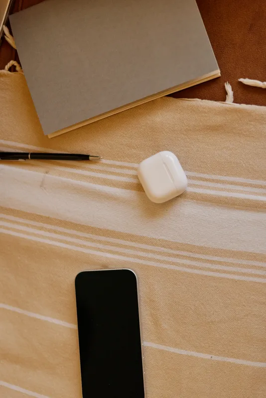 A calm home workspace shows a notebook, pen, and smartphone on a striped beige surface, with a wireless earbuds case nearby, suggesting daily planning and personal productivity at home.
