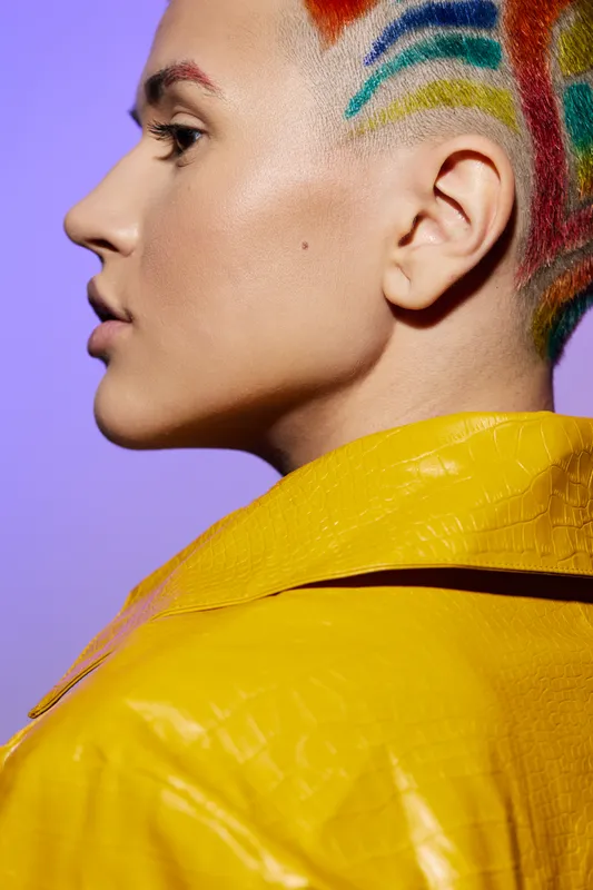 Close-up profile of a woman with colorful rainbow shaved hair and smooth skin wearing a bright yellow textured jacket against a purple background, stylish and modern.