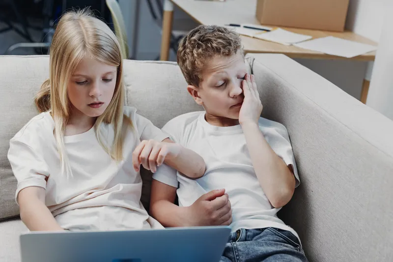 Two children sit on a couch looking tired and distracted, one rubbing eyes while the other holds a laptop. Concept of fatigue and digital distraction.