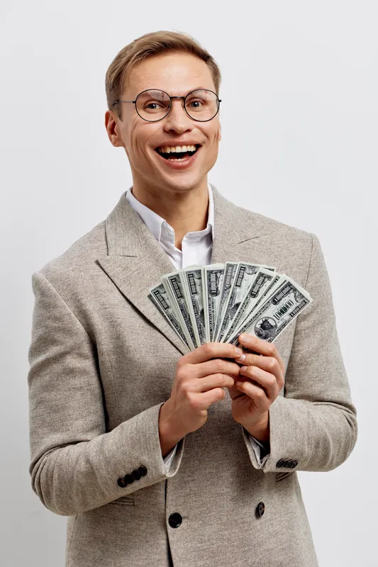 Confident young caucasian man with glasses wearing beige blazer holding a fan of hundred dollar bills smiling happily on light gray background studio portrait. People success concept with money.