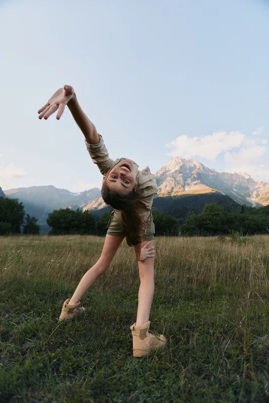 Energetic woman dancing in a grassy field with distant mountains, blue sky, and warm light. Natural, carefree lifestyle and outdoor movement.