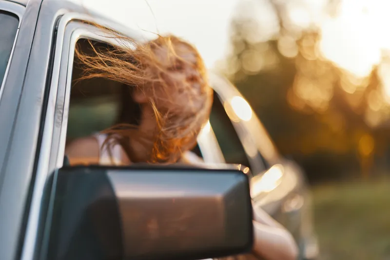 Young woman with flowing hair enjoying a sunny road trip, leaning out of a car window, with a blurred nature background emphasizing freedom and adventure.
