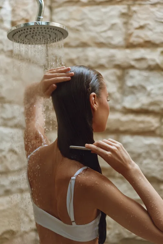 woman combing hair under outdoor shower with stone wall background, wet skin, long dark hair, natural light, summer day, personal care routine