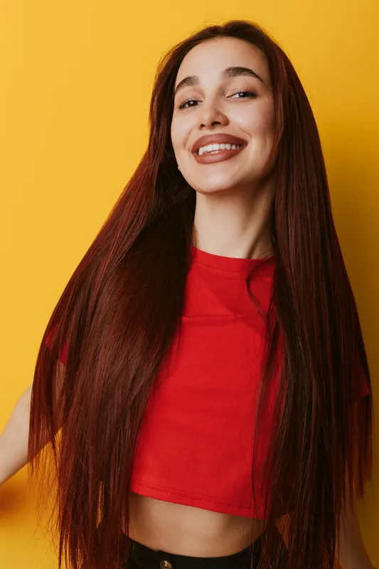 Young woman with long straight hair smiling confidently in casual red top against yellow background. Portrait of happy female with natural makeup and relaxed pose indoors.