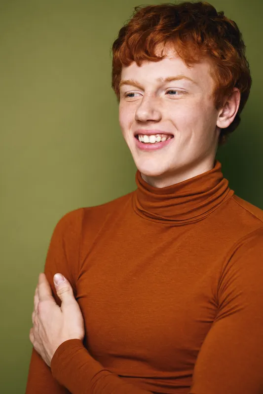 Young man with red hair wearing brown turtleneck smiling and looking aside on olive green background in casual style portrait with natural expression.