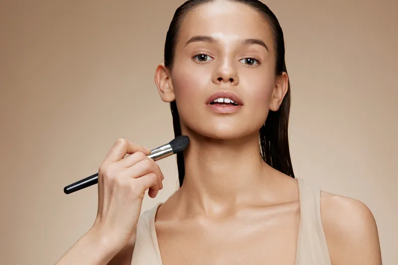 Young model applying makeup with a brush on a neutral background, showcasing natural beauty and elegance, perfect for beauty and skincare concepts