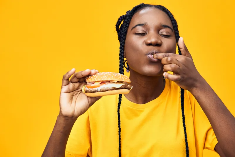 Happy young woman enjoying a delicious hamburger against a bright yellow background, showcasing her love for fast food and vibrant lifestyle