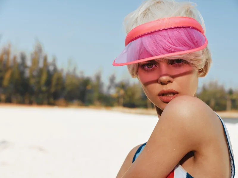 A close up portrait of a young woman on a sunlit beach, wearing a pink visor and swimsuit, capturing bold summer fashion and edgy, contemporary style.