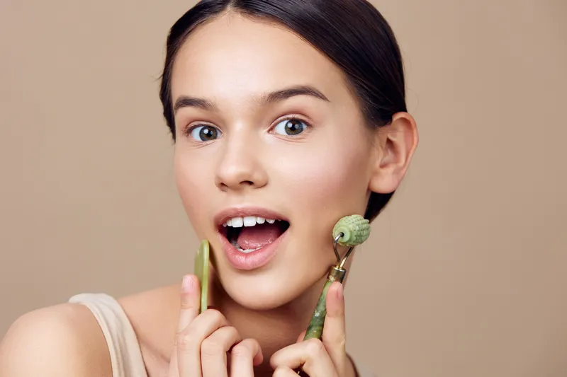 Young woman with radiant skin using jade roller for skincare, showcasing a happy expression against a neutral beige background