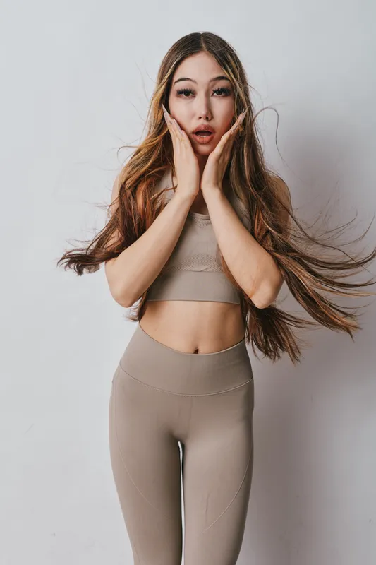 Woman with long hair in beige activewear, standing with hands on cheeks, surprised expression in studio with white background and wind blowing her hair around her face.