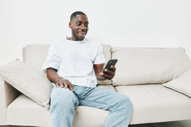 Smiling Black man using smartphone on a casual sofa, dressed in a relaxed outfit, enjoying his time at home in a modern and comfortable setting