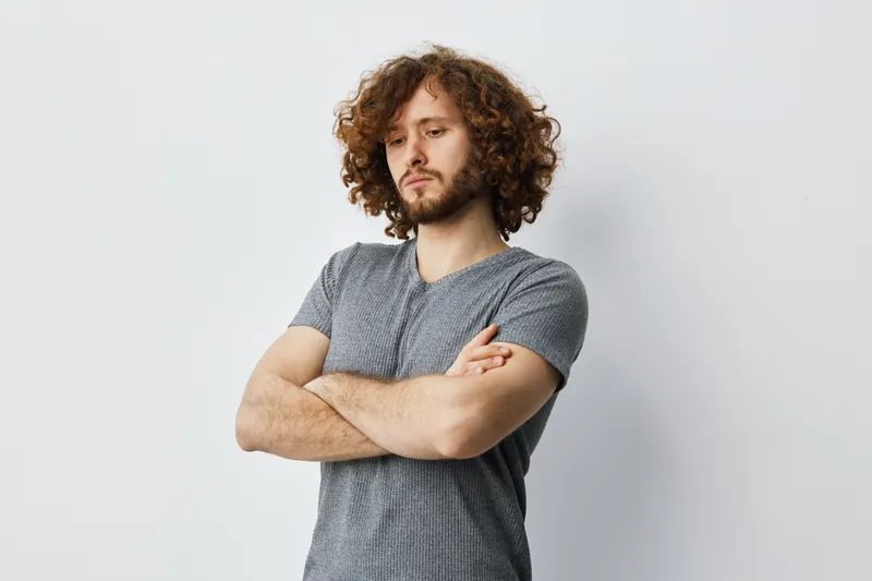 Thoughtful young man with curly hair in a gray t shirt standing against a light background, expressing contemplation and confidence
