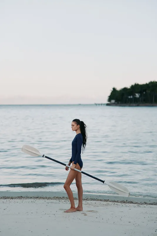 A woman walks along a sandy shoreline carrying a paddle, wearing a navy swimsuit, as calm water and distant trees frame a serene beach scene.