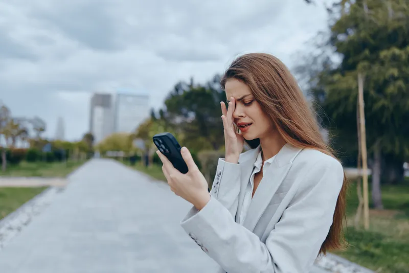 Businesswoman in a blazer experiencing stress while checking her phone, set in an urban park with greenery and modern buildings in the background.