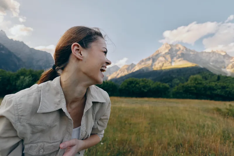 Happy woman enjoys outdoor lifestyle in mountain field smiles broadly looks away wears casual jacket under cloudy sky during daytime.