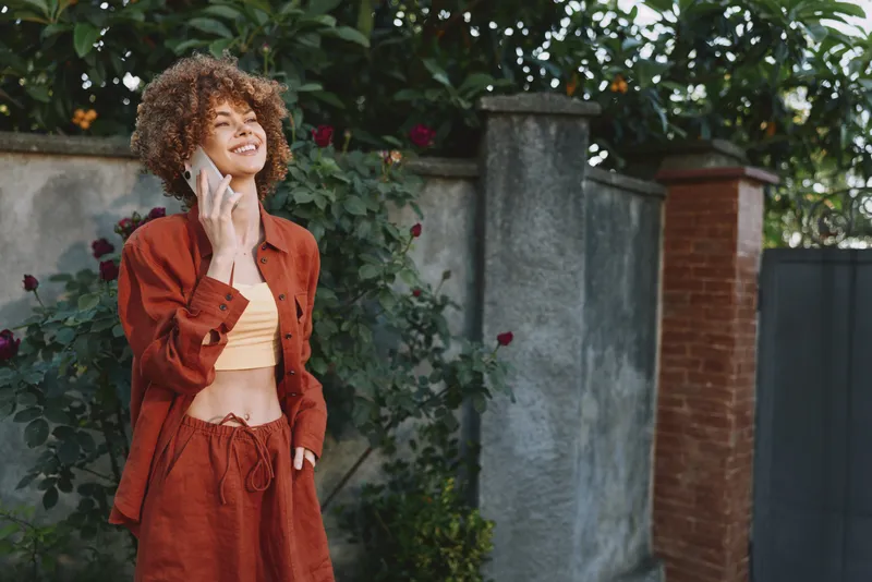 Elegant woman with curly hair wears a red outfit outdoors, smiling beside a stone wall and lush greenery. A lifestyle image highlighting casual fashion, confidence, and everyday joy under bright