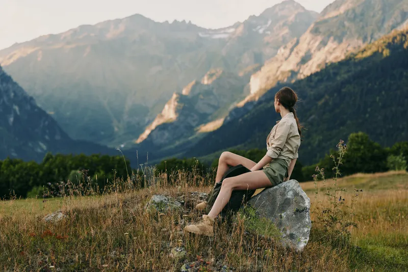 Woman sitting on a rock in a mountain meadow, enjoying outdoor scenery. Natural light highlights the valley and distant peaks, conveying serenity, exploration, and nature connection.
