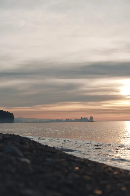 Sunset over calm water by a rocky shore with distant skyline silhouettes, soft clouds reflecting warm hues, a tranquil evening scene suitable for nature, travel, and cityscape compositions.