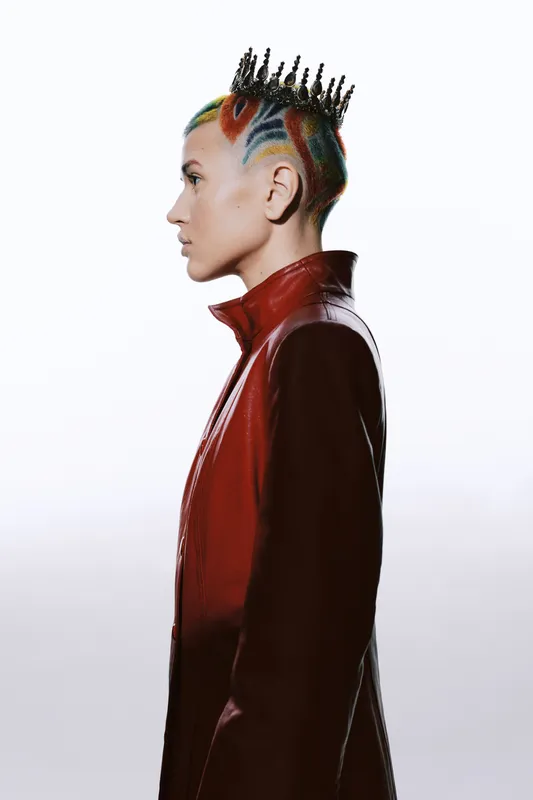 Woman with colorful crown hairstyle wearing a red leather jacket, profile view with a futuristic and edgy fashion style against a clean white background.
