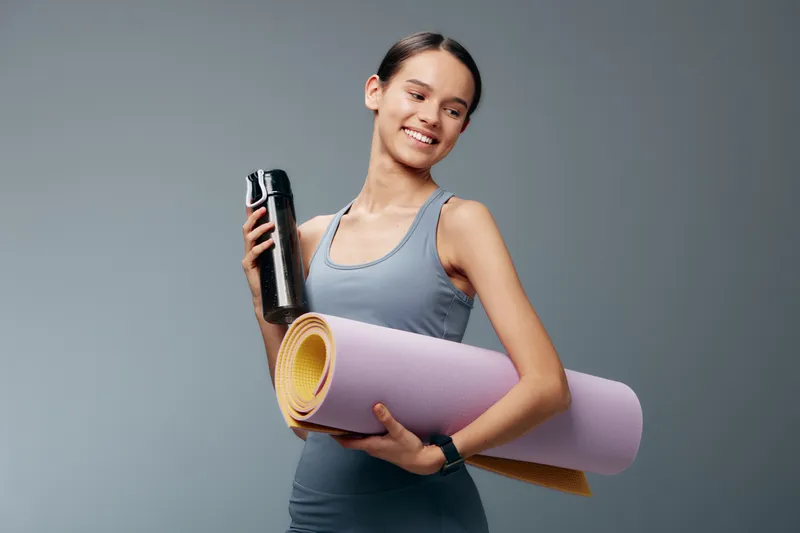 Active woman in athletic wear holds a yoga mat and a bottle, ready for a high energy fitness workout and sport routine in a bright, clean studio with focused mindset and motivation for progress.